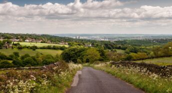 A country lane descends from the Peak District hills into the Mayfield Valley and the suburbs of Sheffield, with the city centre cityscape in the dist
