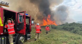 Firefighters at the scene of a wildfire. There are flames in the background, several fire crew and an engine in the foreground