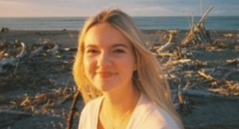 Image of Trustee Emily Johnson. Image shows the head and shoulders of a girl in her early twenties. She is wearing a white cardigan and has blonde hair and is smiling. Int he background is a beach with beach and sea debrie across the sand. You can see the sky disappearing into the horizon with light cloud cover